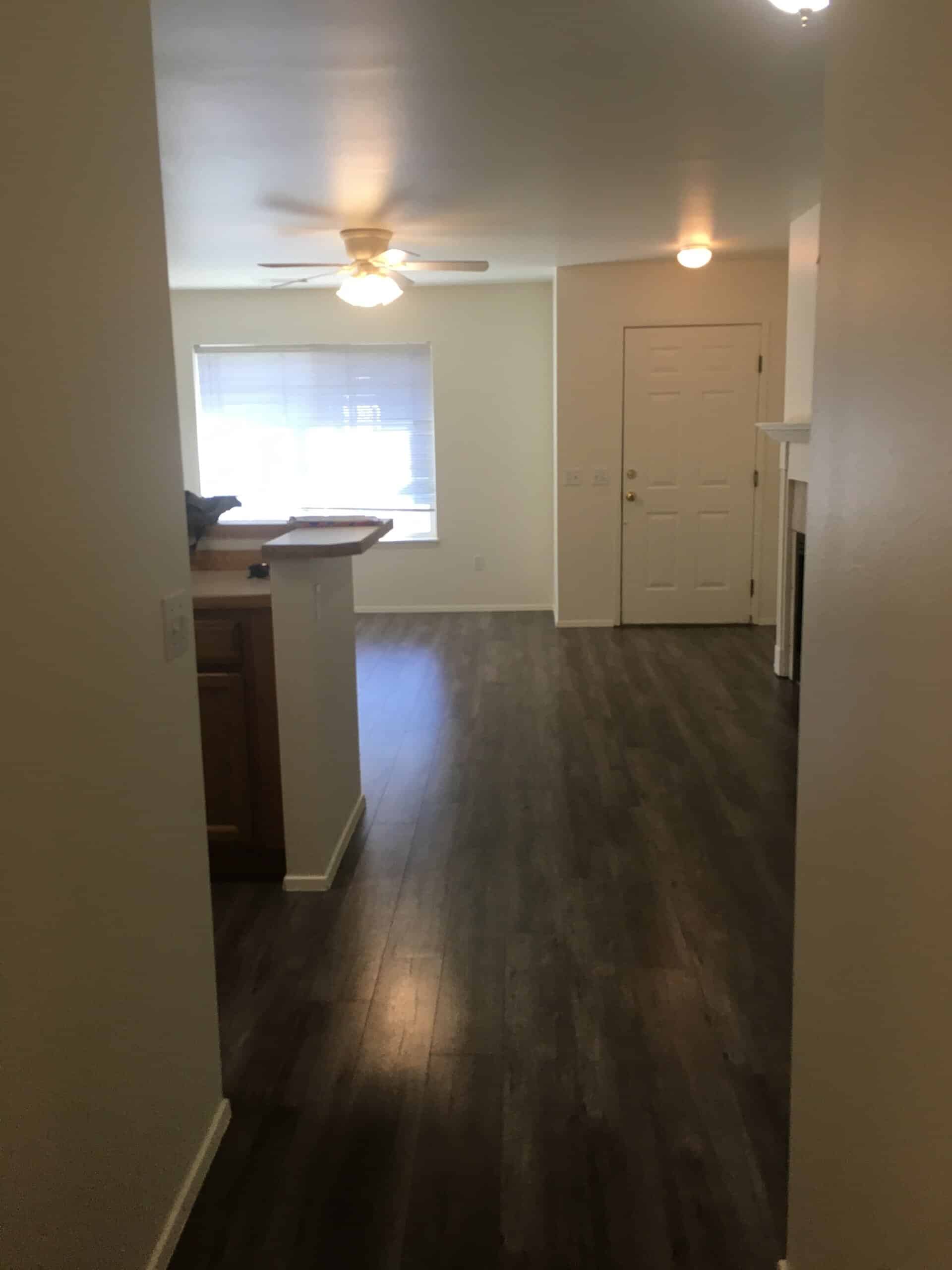 A room with dark plank flooring, a window with blinds, a ceiling fan with a light, a closed door, and a counter.