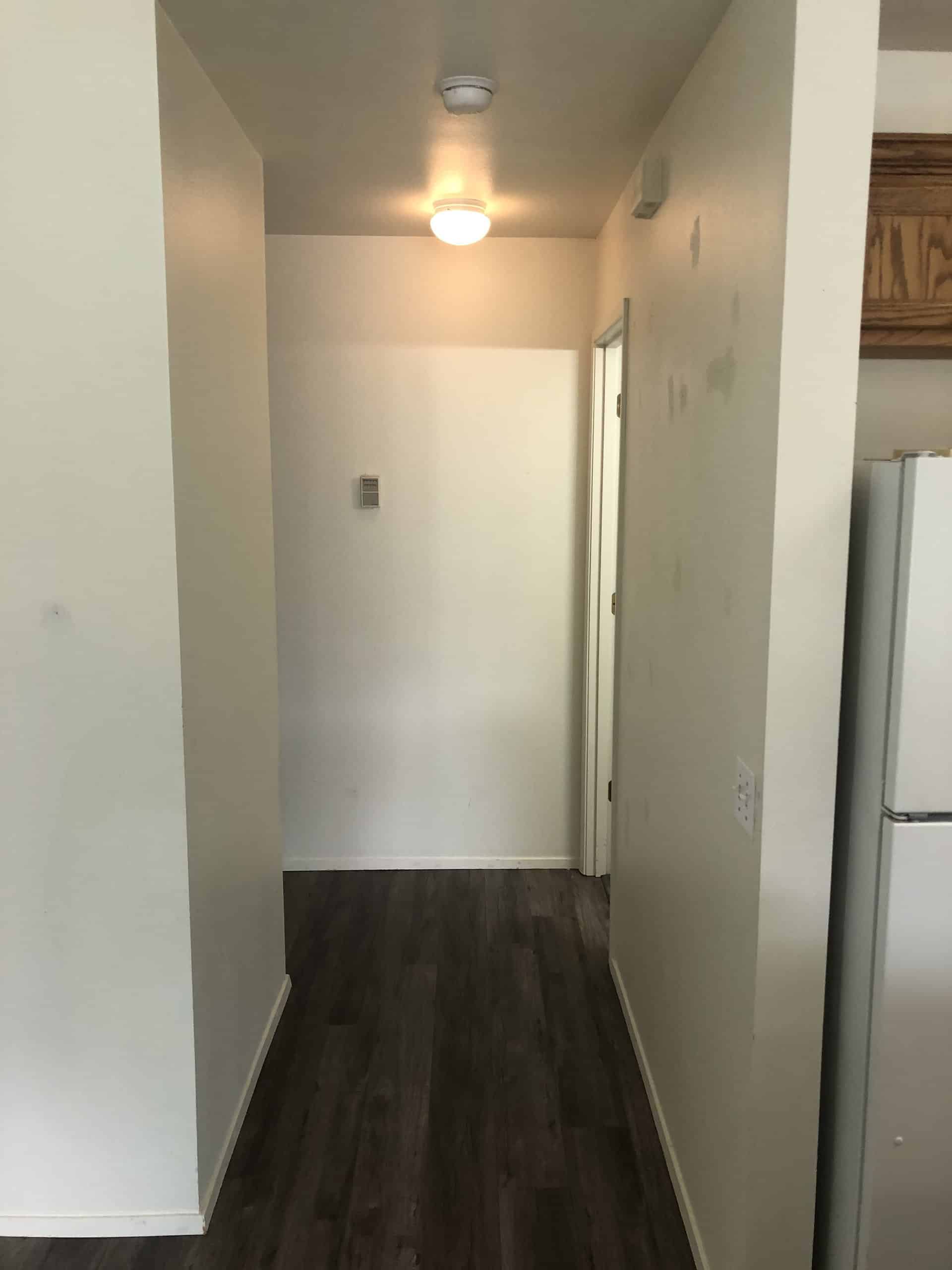 A narrow hallway with dark wood flooring, light walls, a lit ceiling fixture, and a doorway on the right.