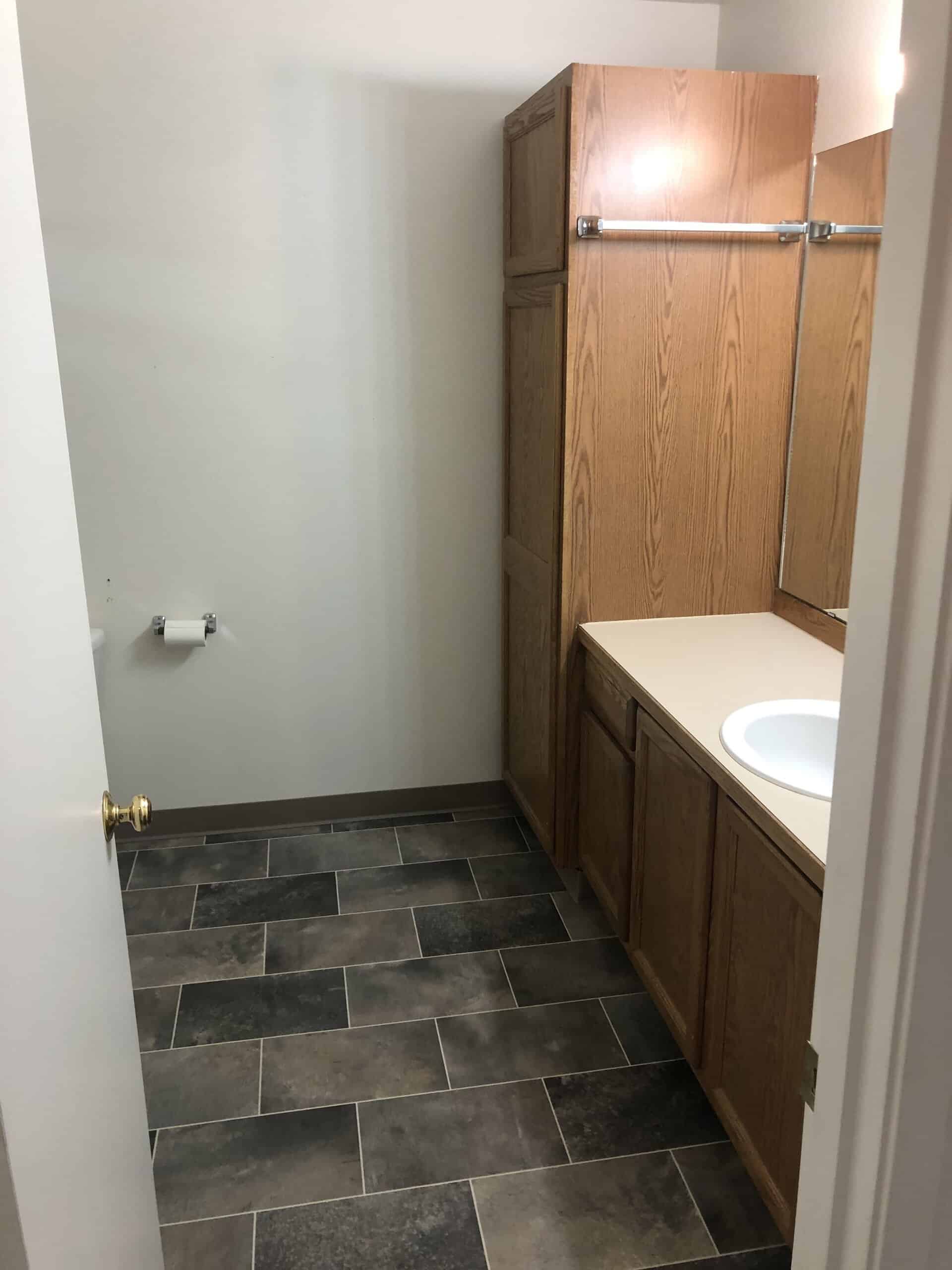 A bathroom with a wood vanity, white sink, wall mirror, dark tiled floor, and toilet paper holder.