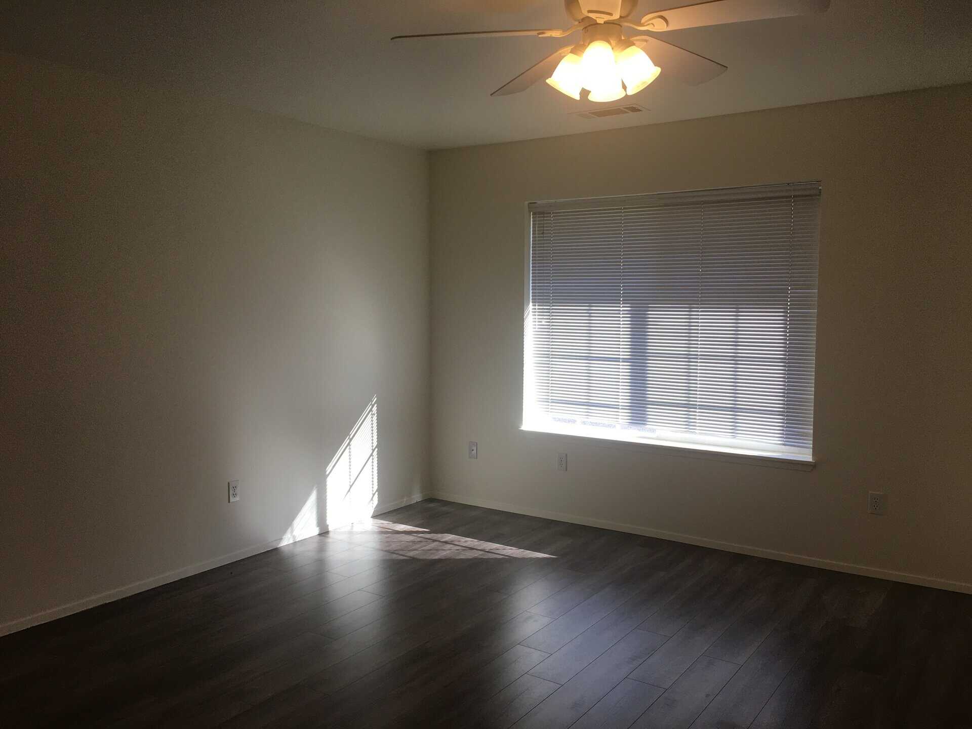 An empty room has dark plank flooring, a window with horizontal blinds, and a lit ceiling fan.