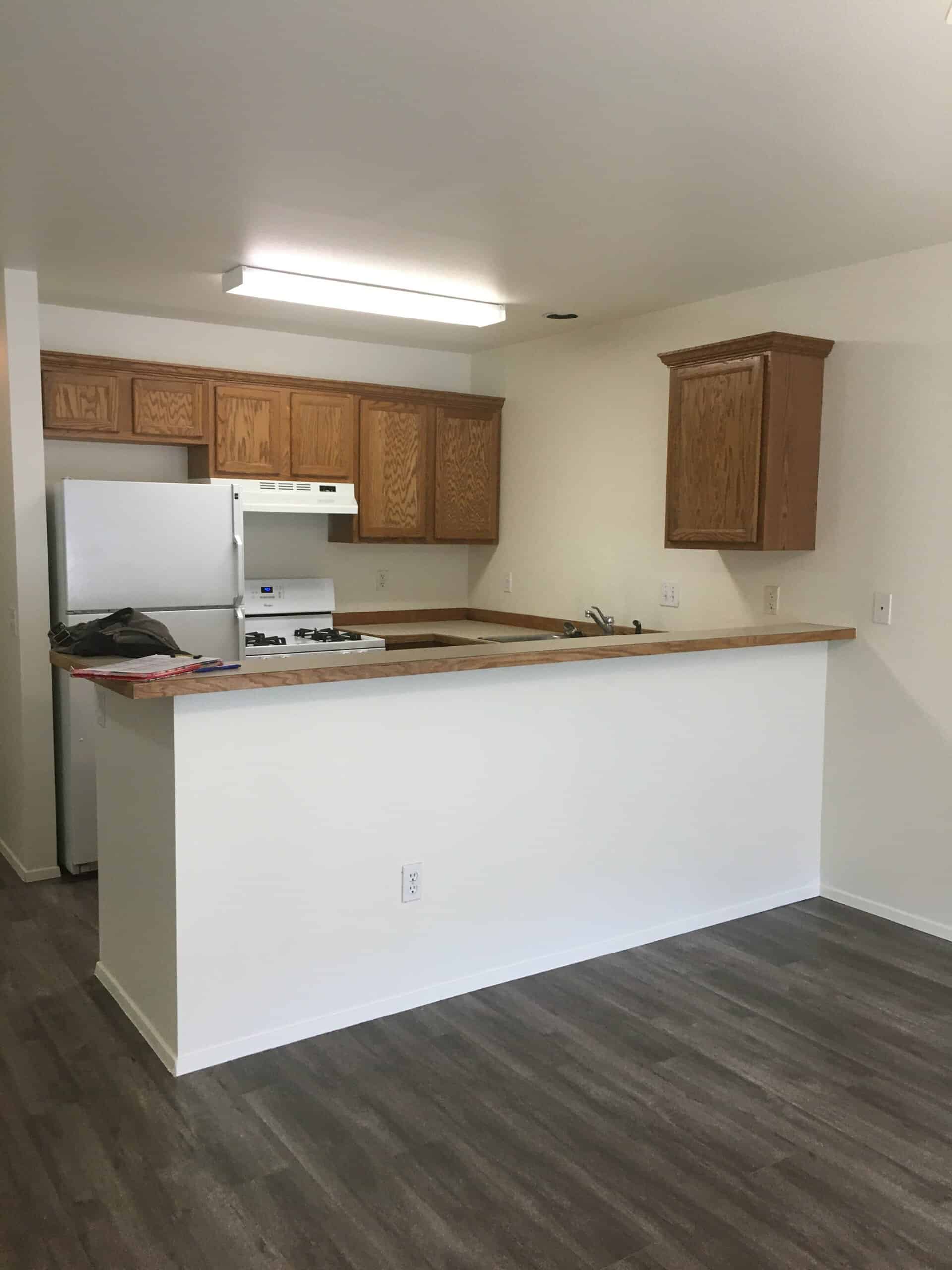 A kitchen with wood cabinets, a white refrigerator and stove, a sink, and a wood-topped peninsula above dark flooring.