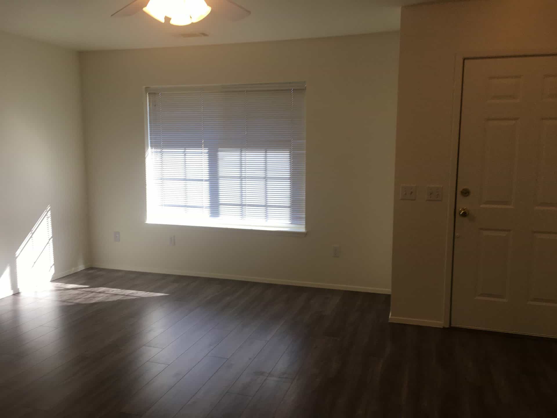 An empty room has dark plank flooring, a window with blinds, a ceiling fan and light, and a paneled door.