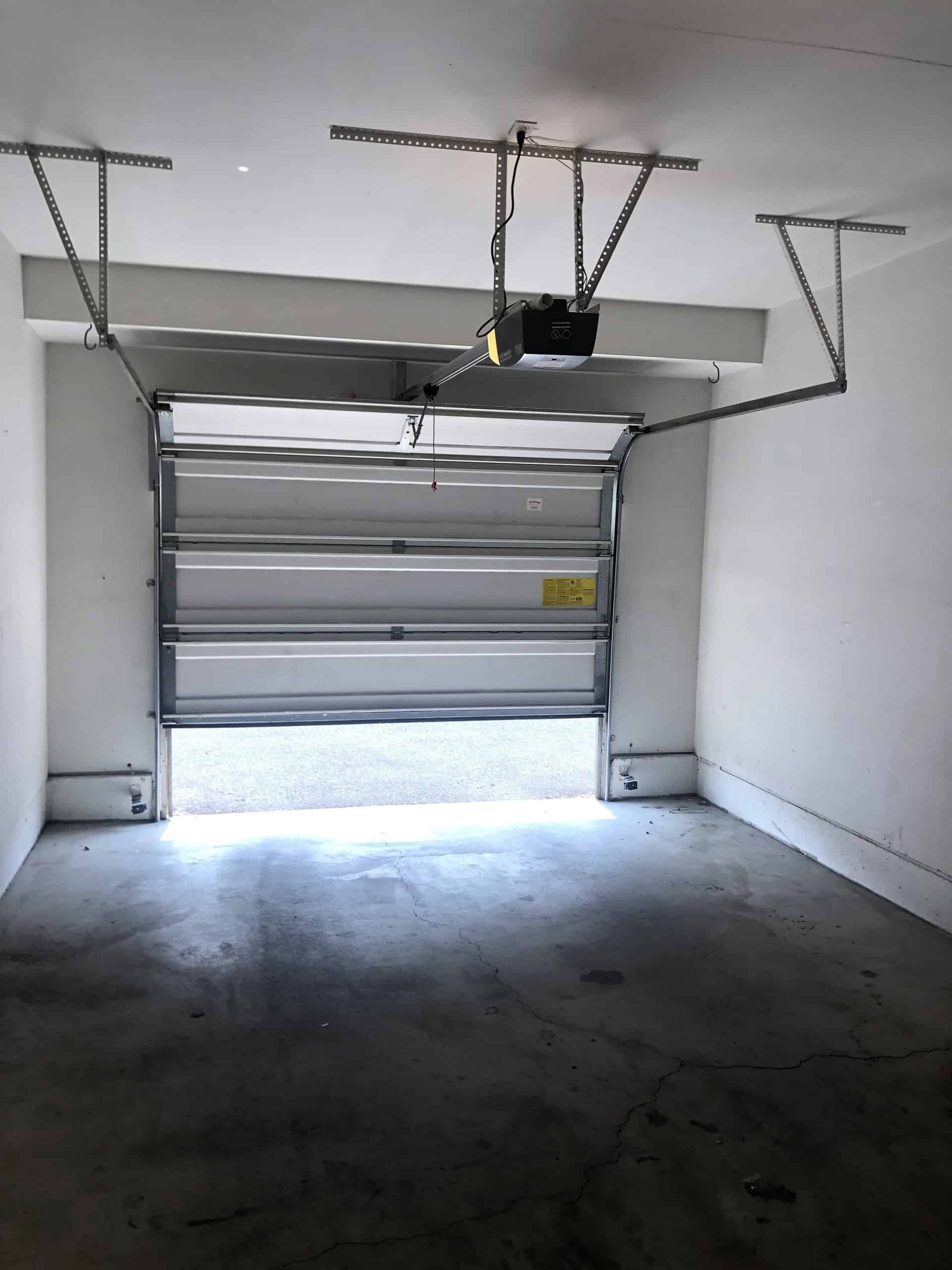 A segmented garage door is partly open, with its ceiling-mounted opener and metal tracks visible above a cracked concrete floor.