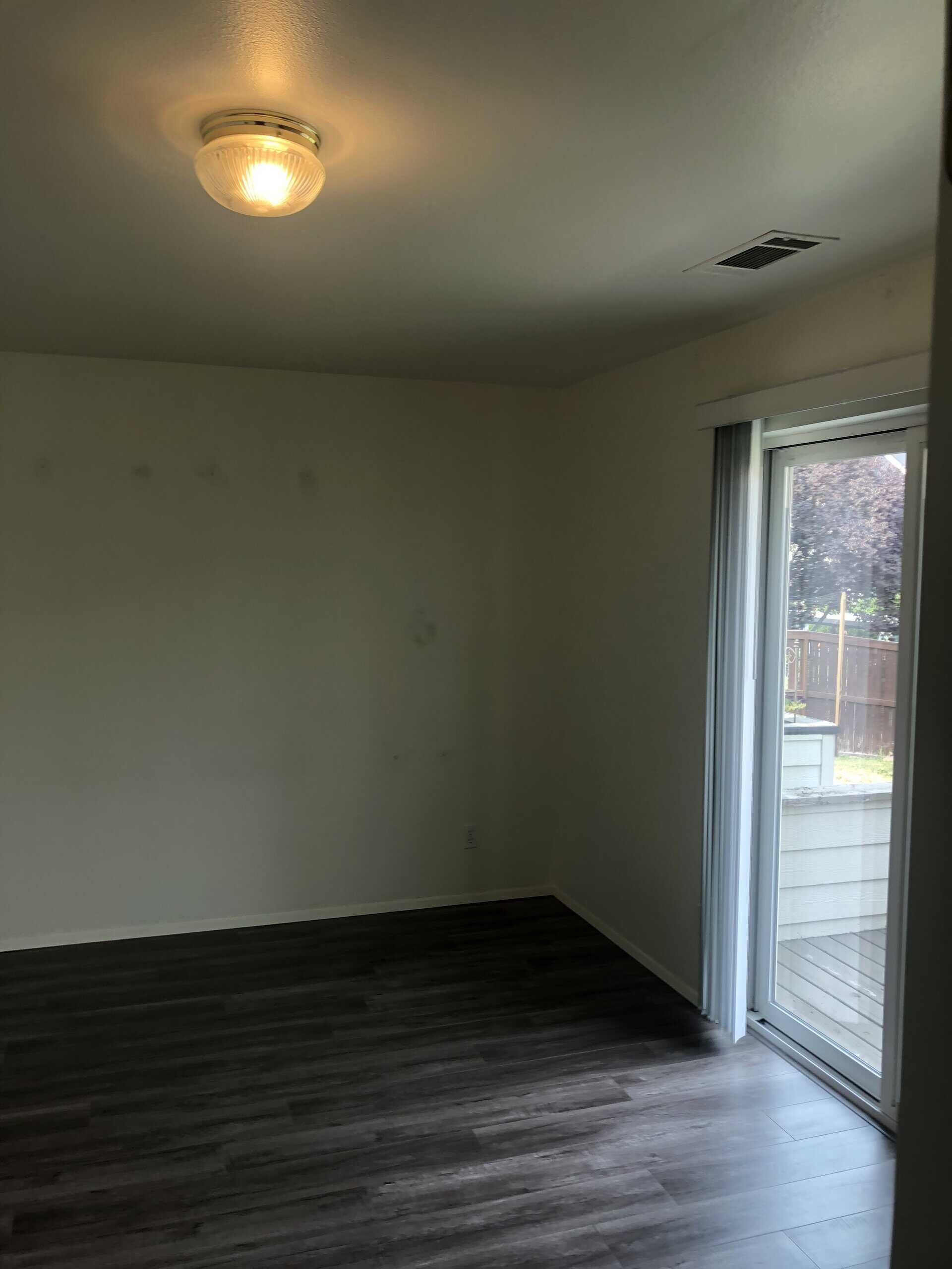 An empty room with dark plank flooring, pale walls, a lit ceiling fixture, and a sliding glass door.