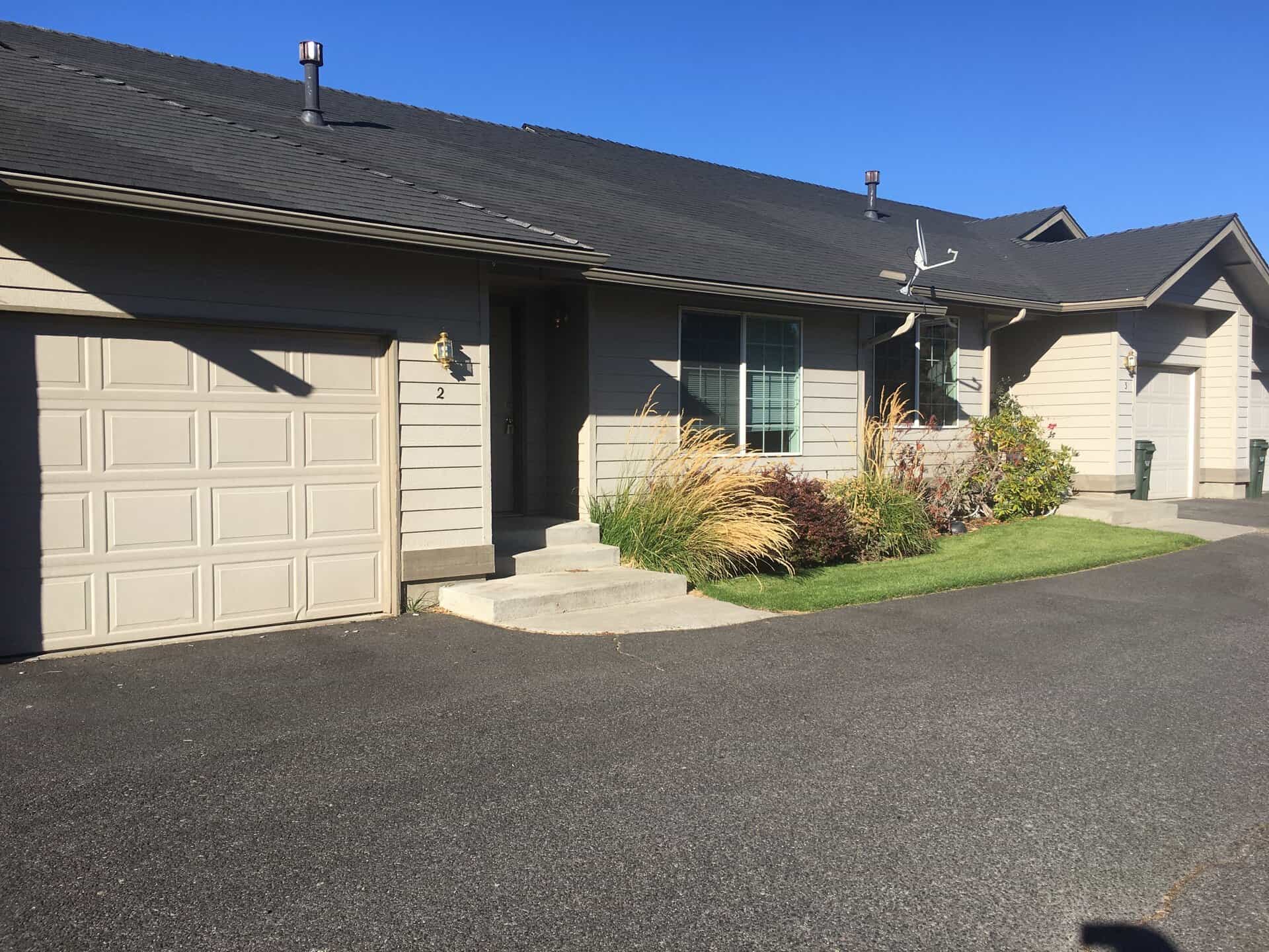 A row of attached homes with dark shingle roofs, garage doors, front windows, entry steps, and planted shrubs beside a paved drive.