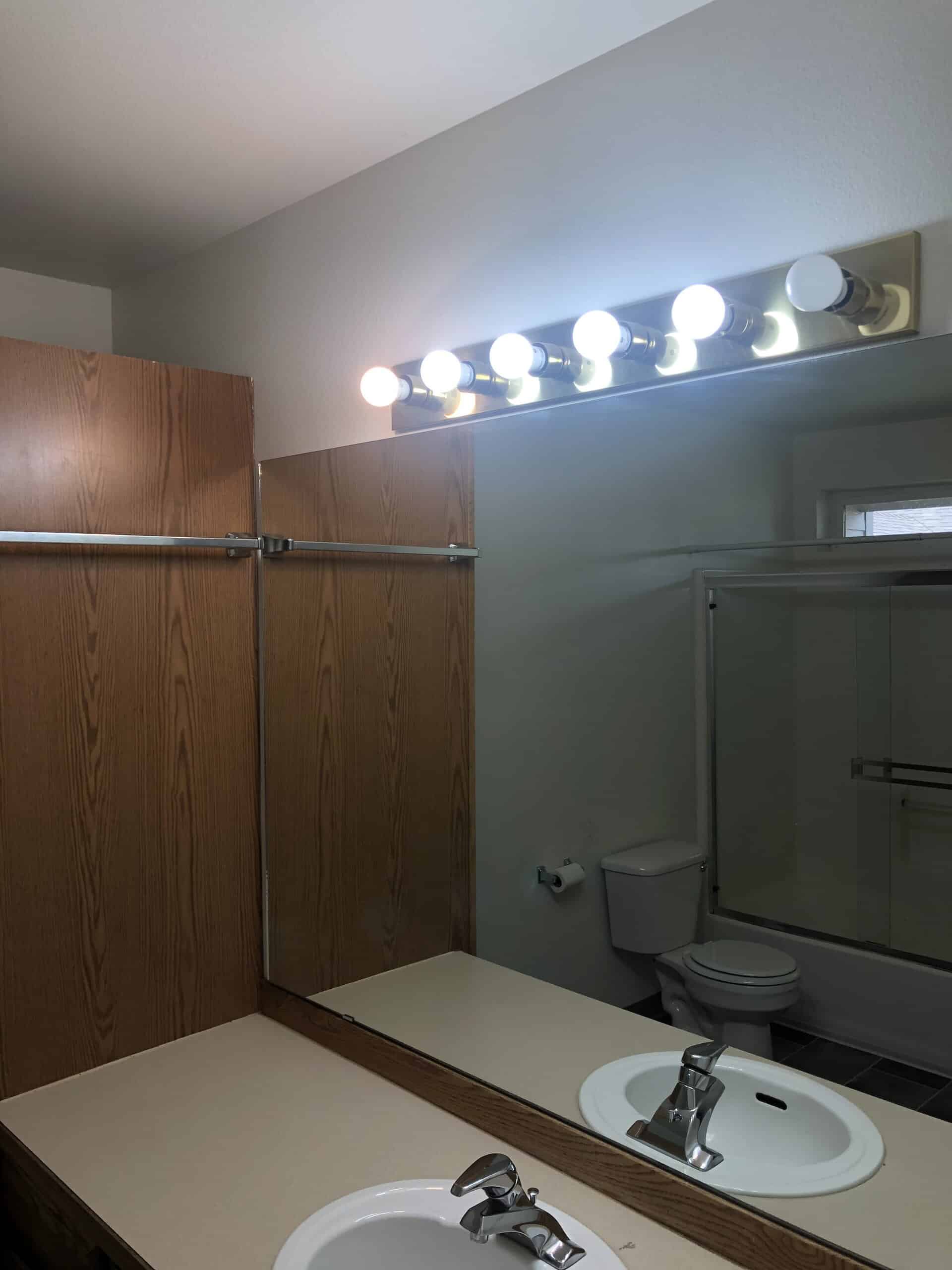 A bathroom vanity with a white sink, large mirror, wood-grain cabinets, and a row of lights above.