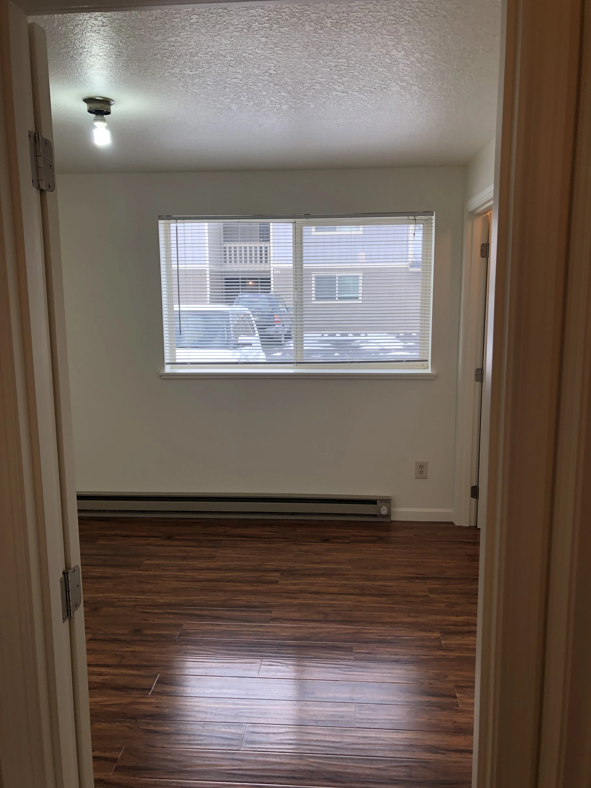 An empty room with dark wood-pattern flooring, a wide window with horizontal blinds, and a long heater beneath it.