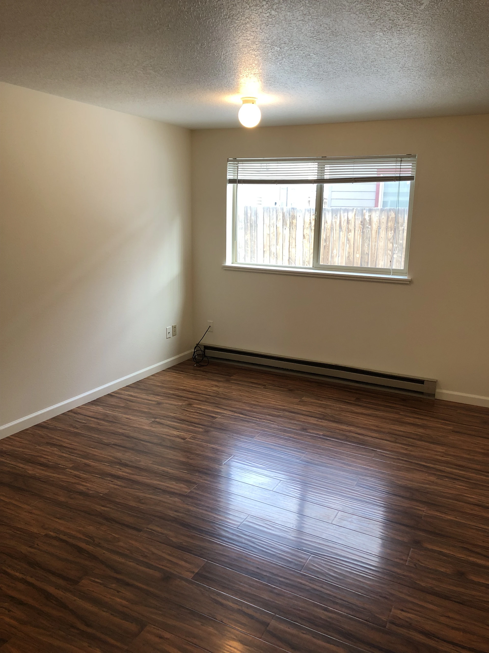 An empty room with brown plank flooring, beige walls, a window with blinds, a ceiling light, and a baseboard heater.