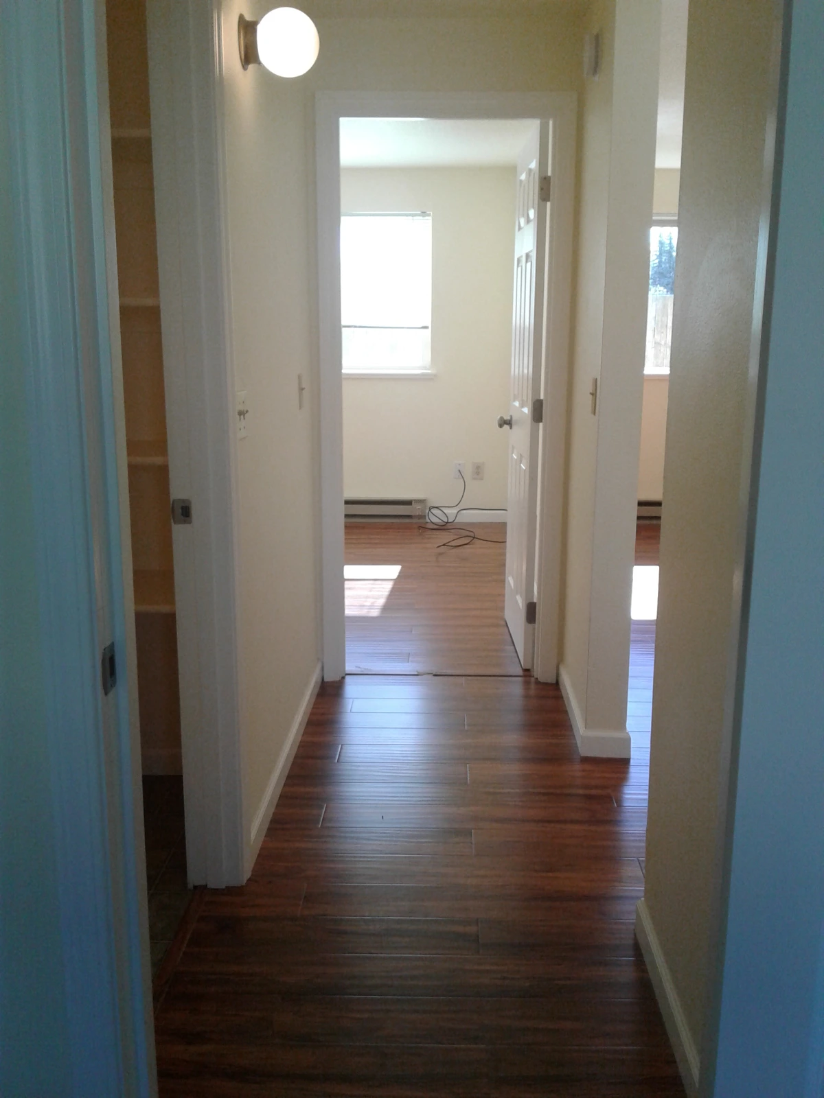 A narrow hallway with dark wood flooring leads to a room with a window; an open door and wall sconce are visible.