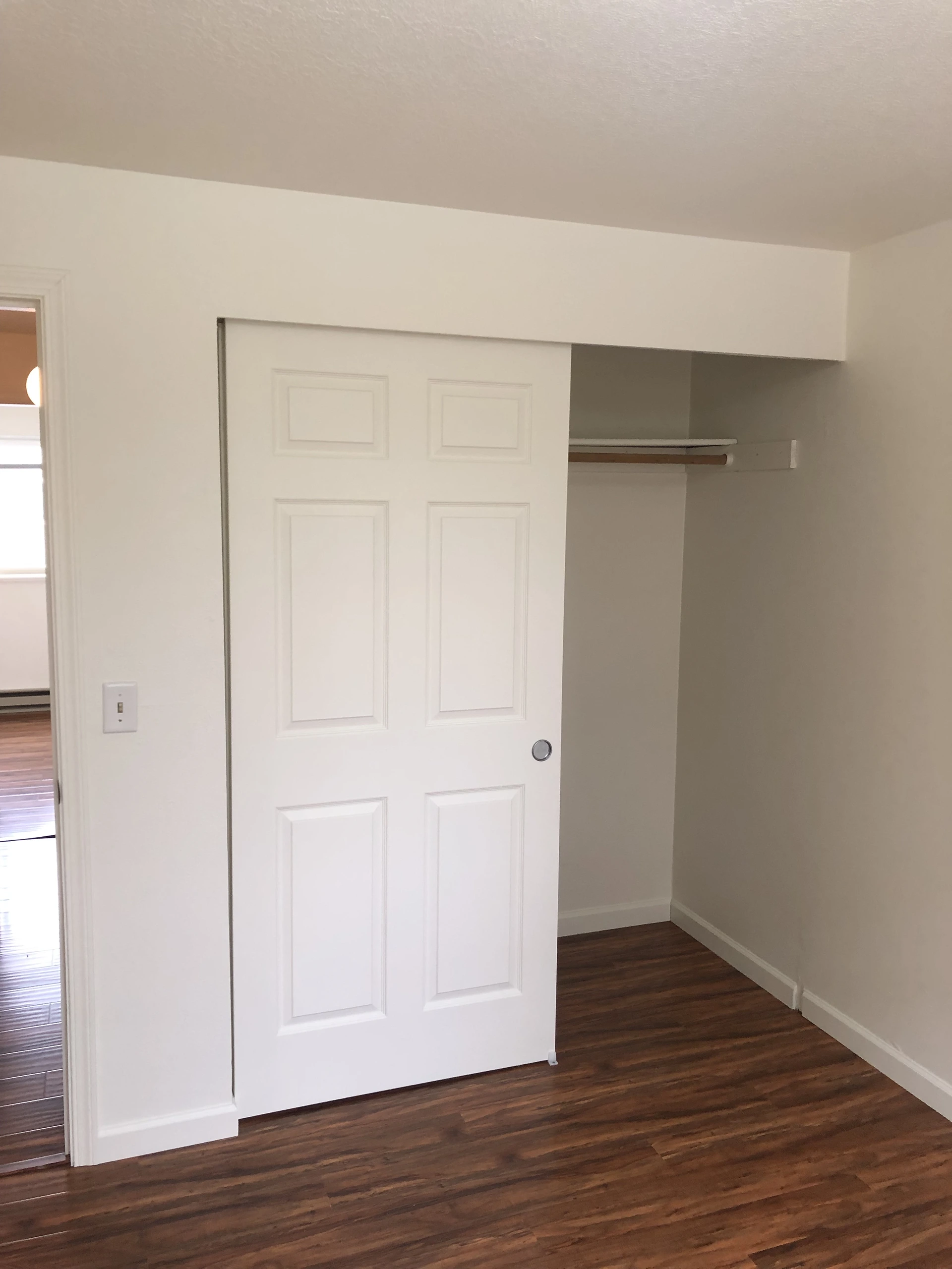 A white sliding paneled door partly covers a closet with a shelf and hanging rod above dark wood-patterned flooring.