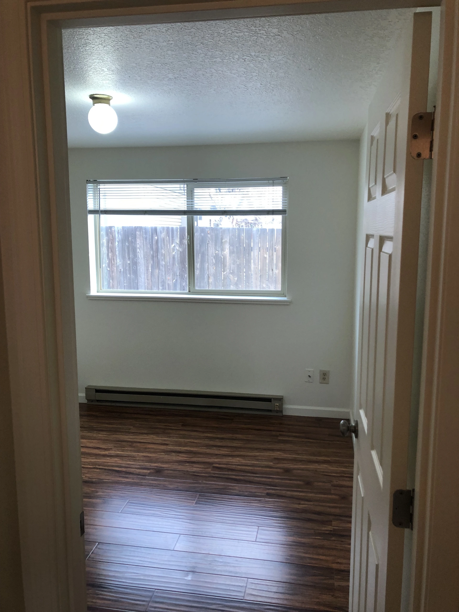 An empty room with dark wood-patterned flooring, a window with blinds, a ceiling light, and a baseboard heater.