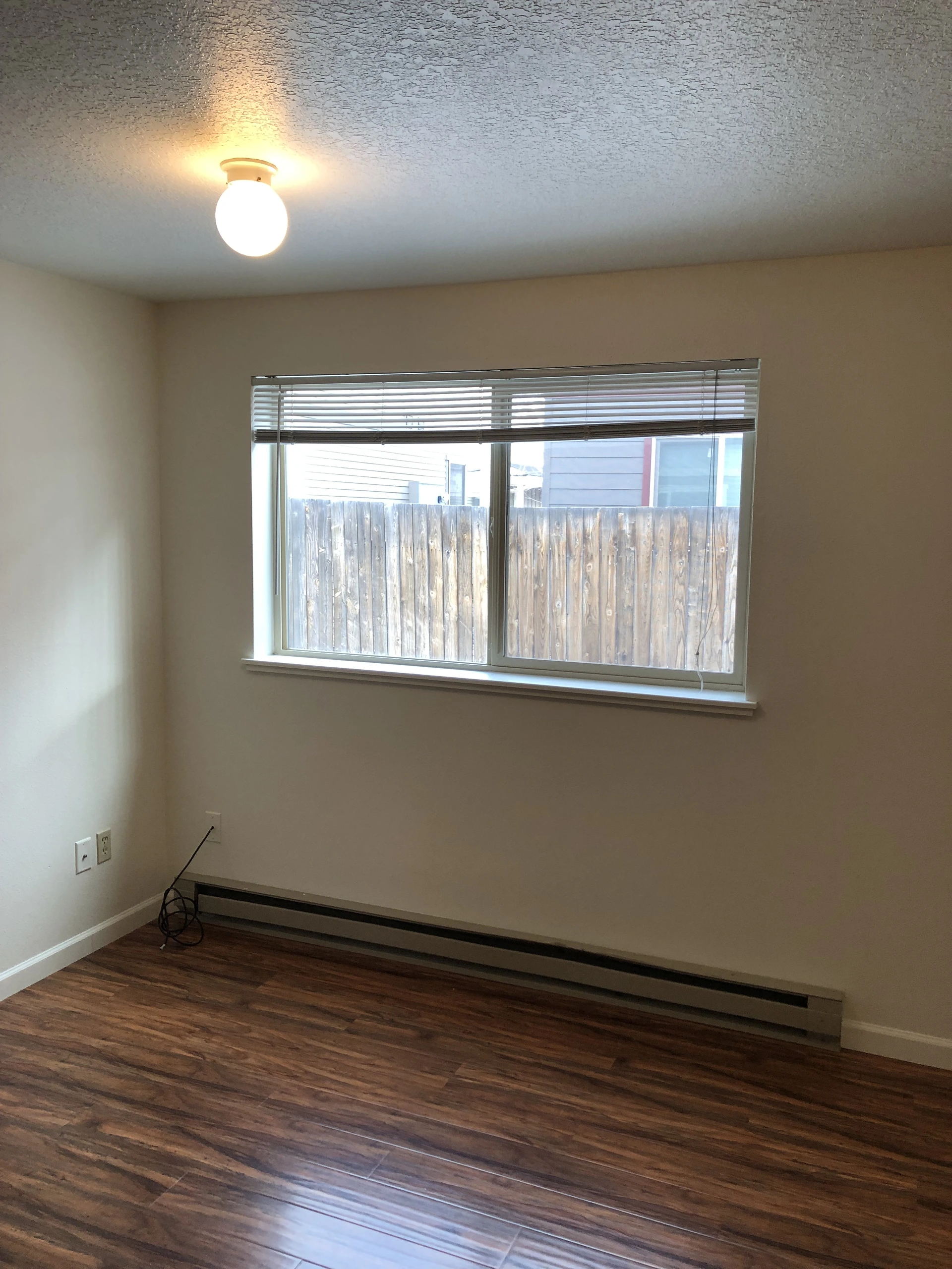 An empty room with a lit ceiling fixture, a window with blinds, a long baseboard heater, and dark wood-patterned flooring.