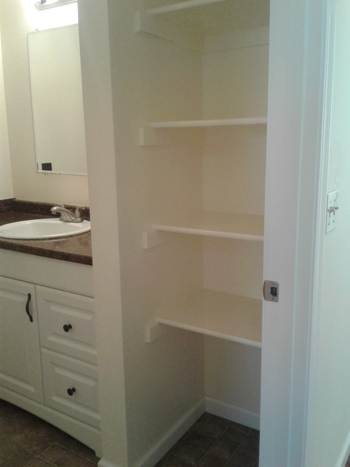 A bathroom vanity with an oval sink and mirror stands beside an open recess with three shelves.