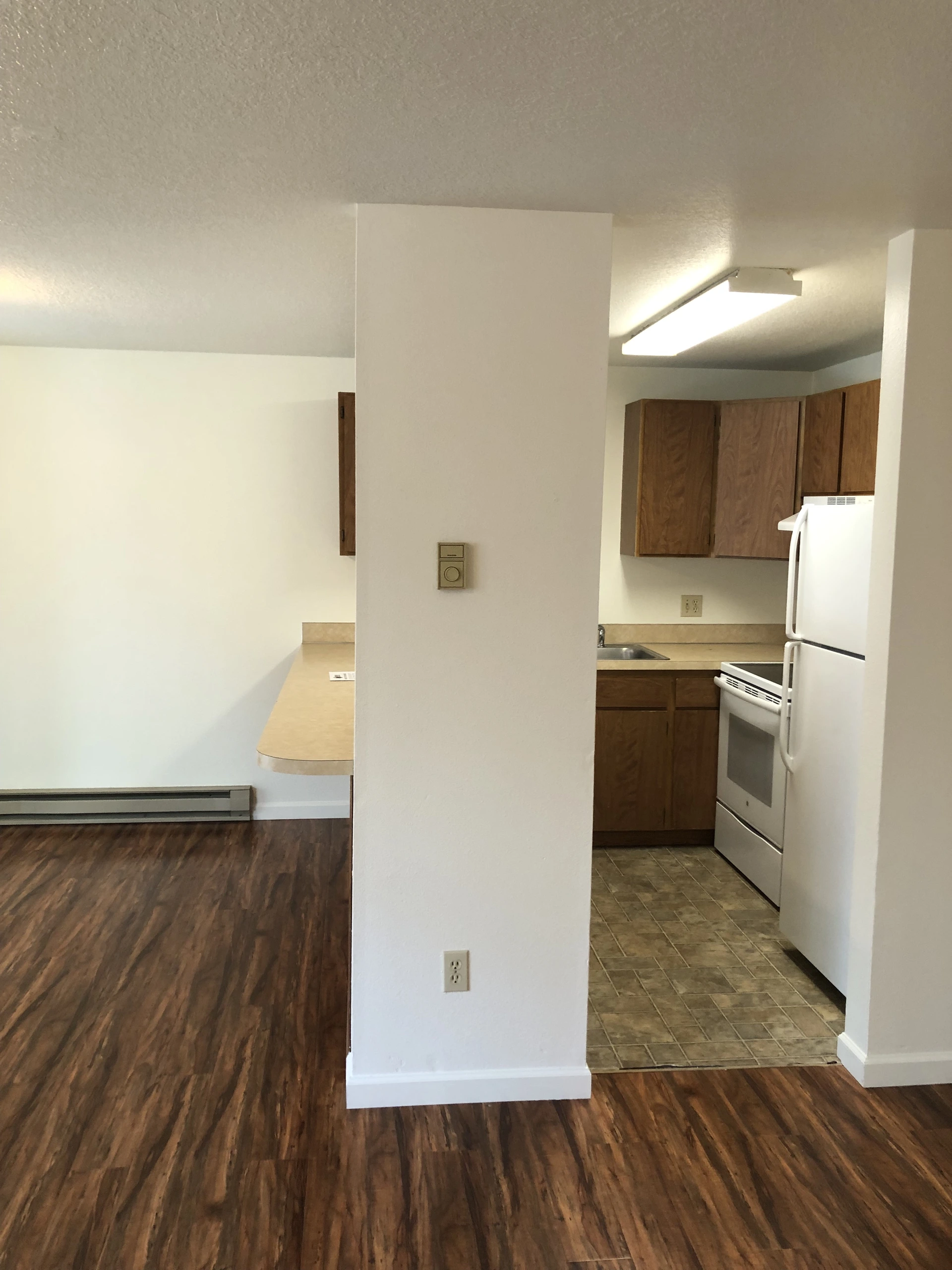 A kitchen with wood cabinets, a sink, white refrigerator and stove sits beside a room with dark wood flooring.