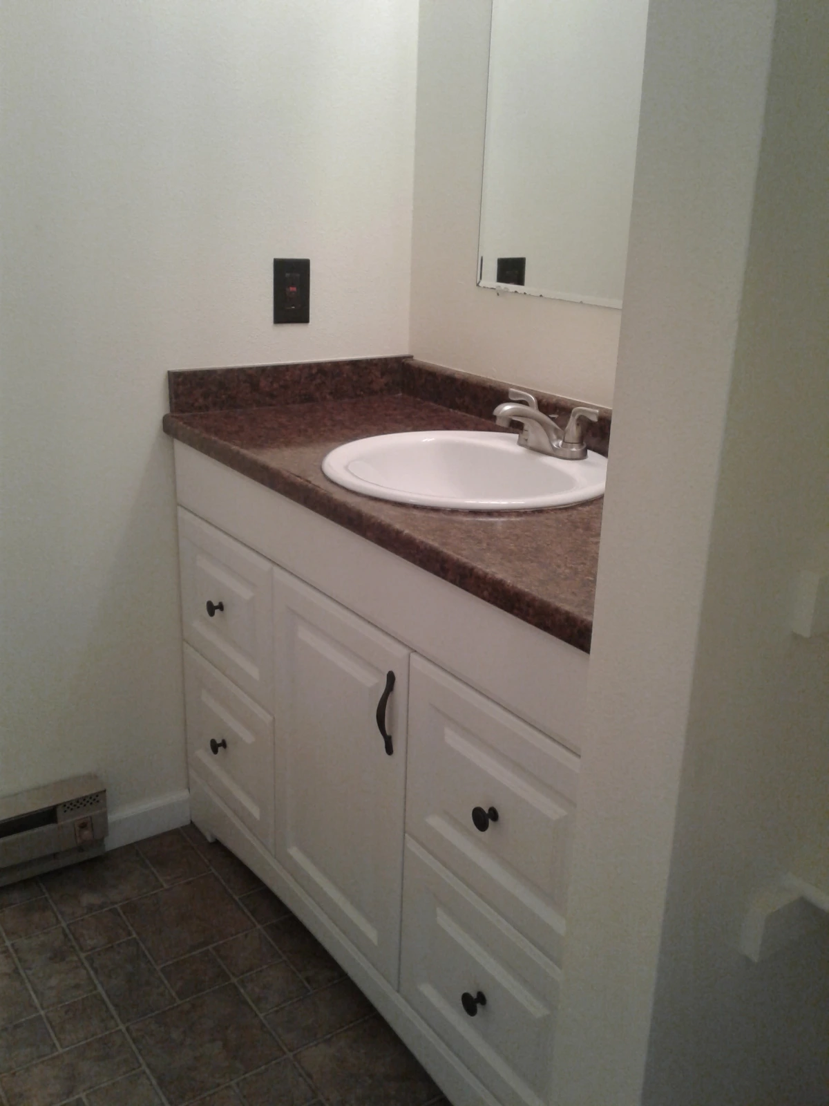 A white sink sits in a brown countertop above white cabinets, with a mirror on the wall and tiled floor below.